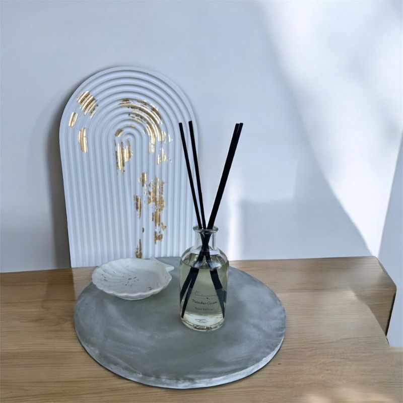 Σύνθεση από στρογγυλό τσιμεντένιο χειροποίητου δίσκος με εφέ μαρμάρου σε λαδί απόχρωση, λευκό κοχύλι, reed diffuser σε γυάλινο δοχείο, και τσιμεντένια αψίδα με φύλλα χρυσού από τσιμέντο.